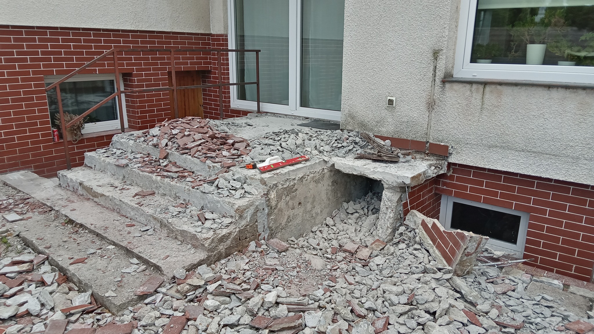 Demontaż schodów wejściowych z czerwonej cegły i betonu, gruz na ziemi, poziomica leży na krawędzi, widoczne okno piwniczne i drzwi wejściowe.