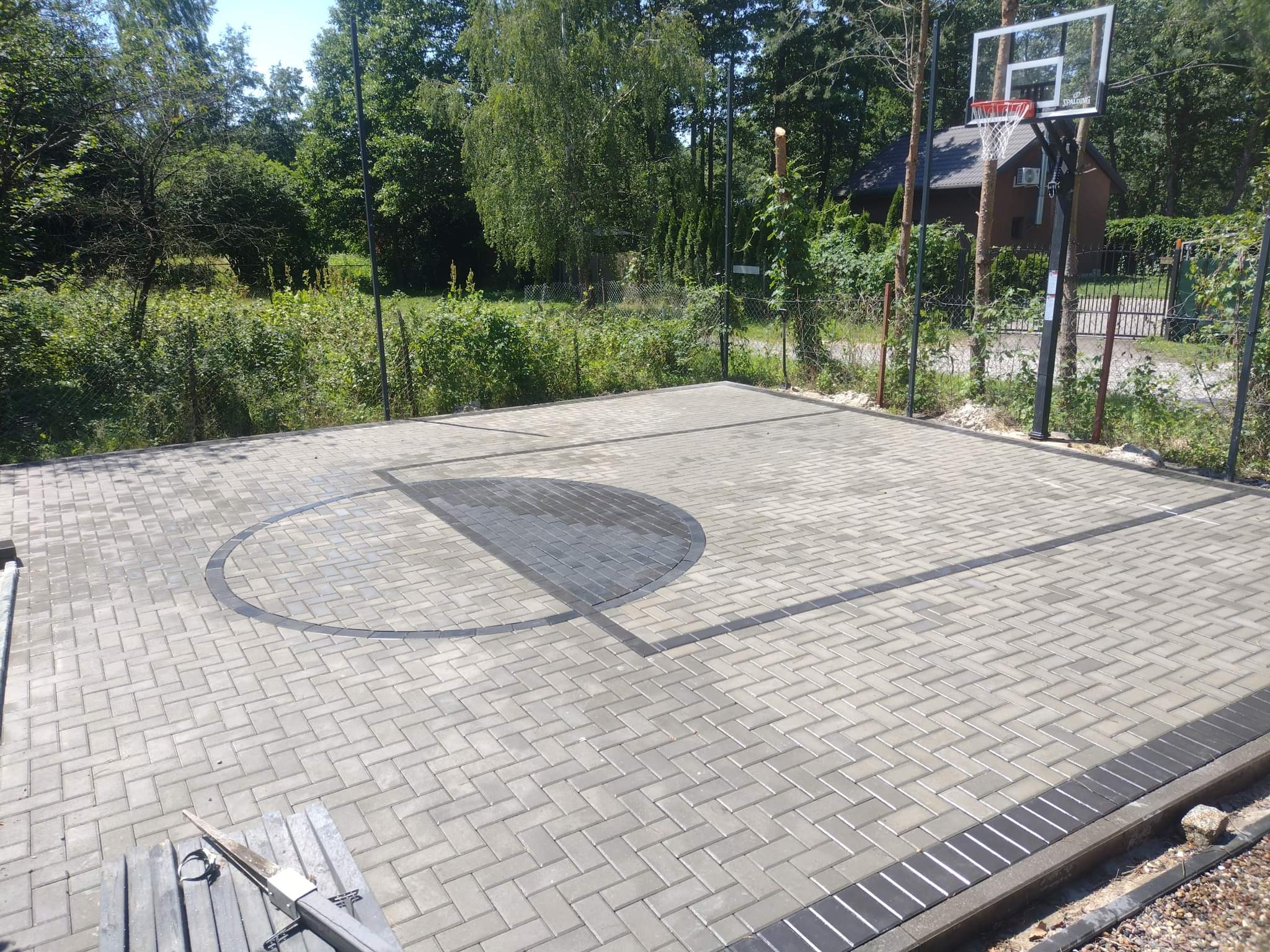 Wykonane z jasnoszarej kostki betonowej boisko do koszykówki z wyznaczonymi liniami i kołem środkowym z ciemniejszej kostki, otoczone zielenią i ogrodzeniem z siatki.