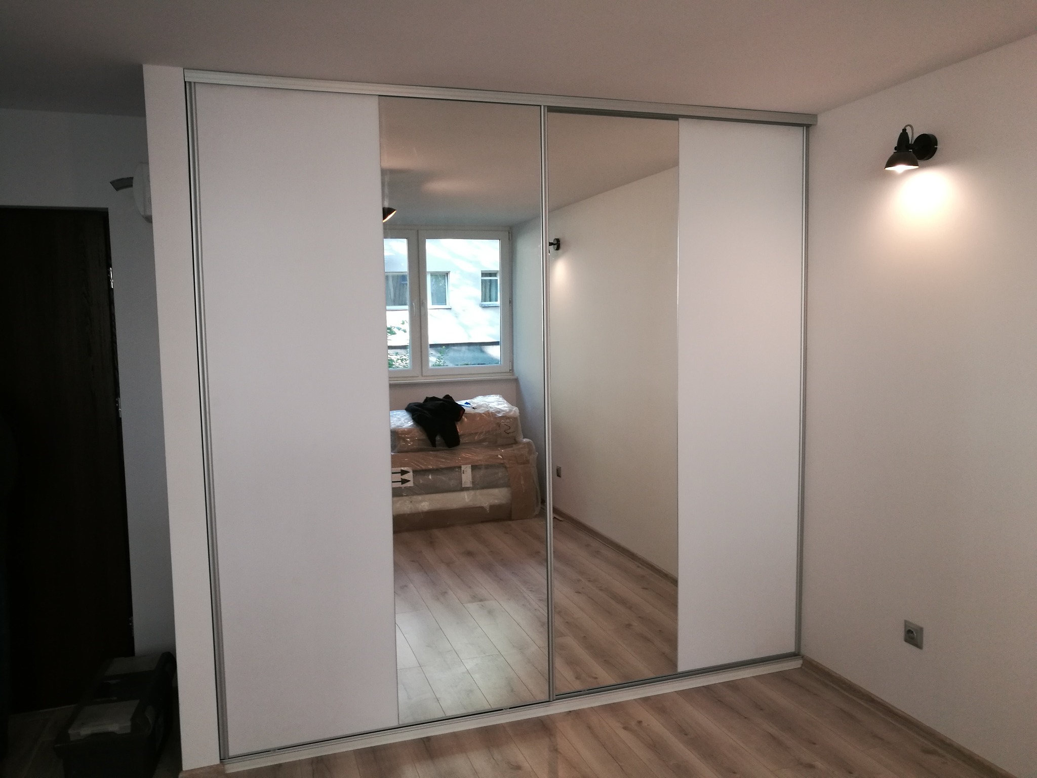 Nowoczesna szafa wnękowa z przesuwnymi drzwiami z białymi panelami i lustrem, odbijająca pokój z oknem i kartonami, oświetlona lampą ścienną.