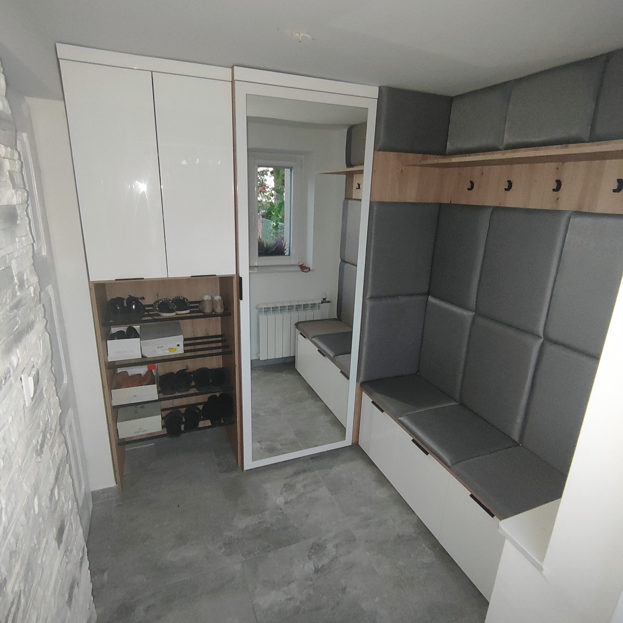 Nowoczesna aranżacja przedpokoju z białą szafą, lustrem i siedziskiem z szarym obiciem, widoczna półka na buty i wieszaki.