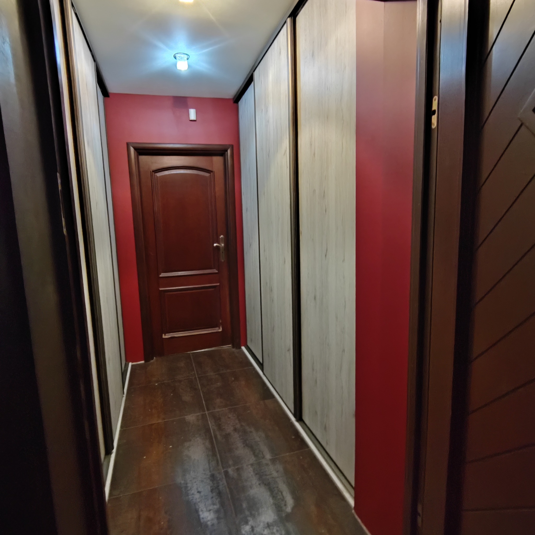 Wnętrze korytarza z zabudowaną szafą przesuwną z drewnopodobnymi frontami, ciemnoczerwonymi ścianami i brązowymi drzwiami.