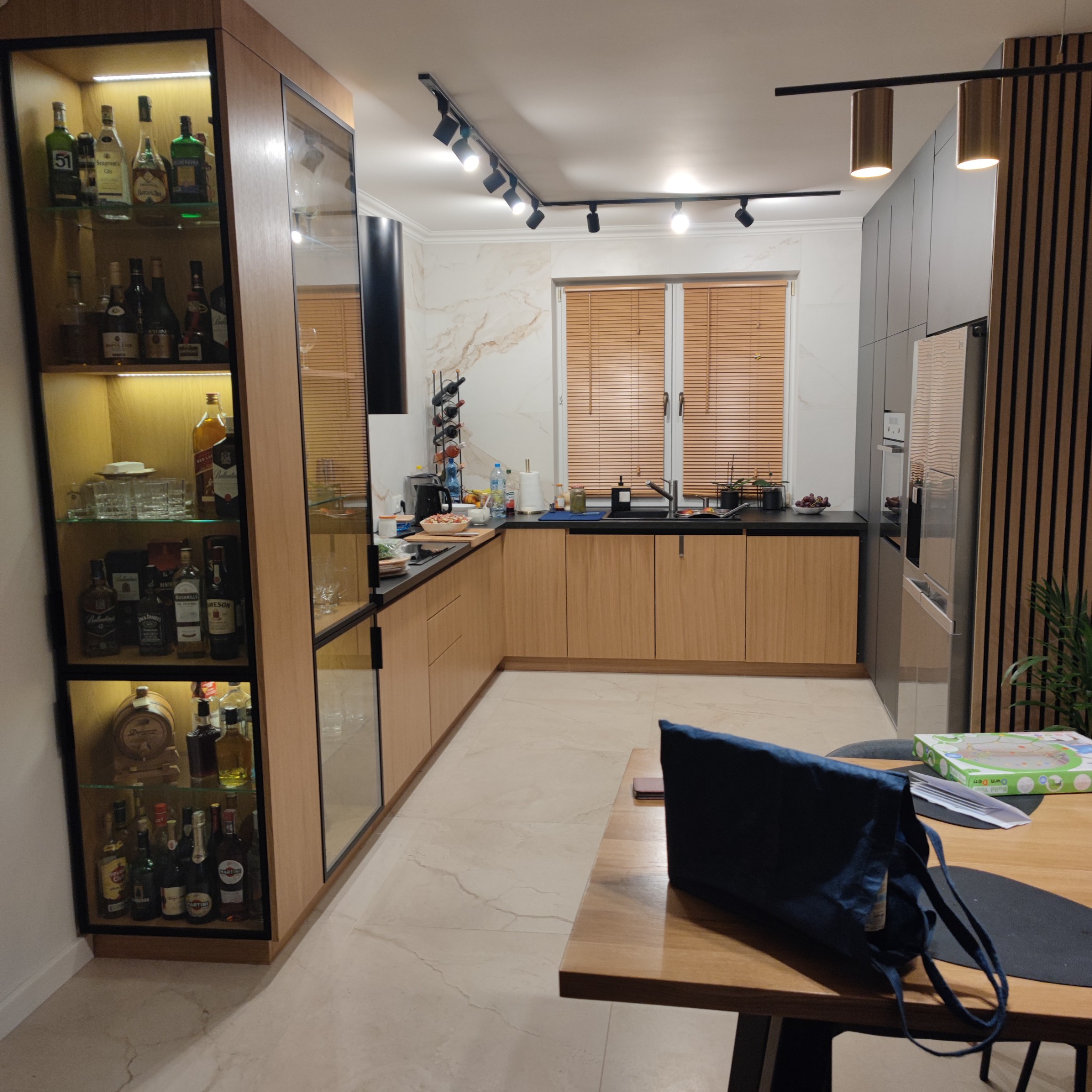 Nowoczesna kuchnia z jasnymi drewnianymi frontami, czarnym blatem, szklaną witryną z alkoholem i oświetleniem punktowym.