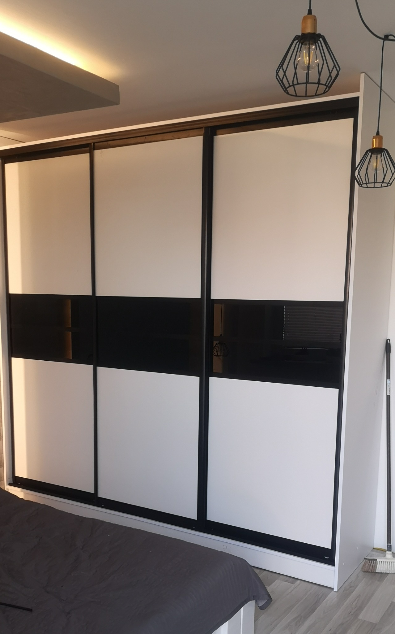 Nowoczesna szafa przesuwna z białymi frontami i czarnym pasem pośrodku, oświetlenie sufitowe i designerskie lampy wiszące w sypialni.