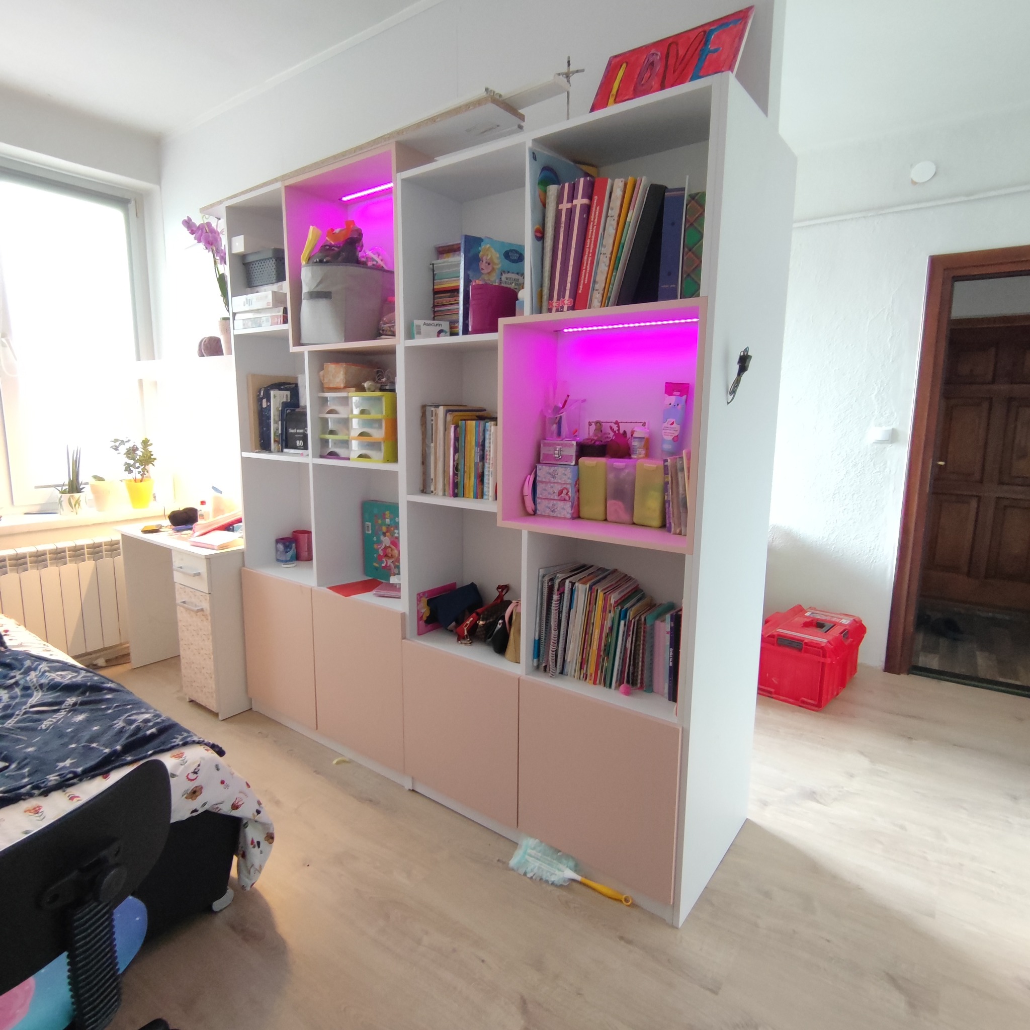 Biały regał z różowymi akcentami, oświetlony fioletowym światłem, oddzielający przestrzeń w pokoju, wypełniony książkami i dekoracjami.