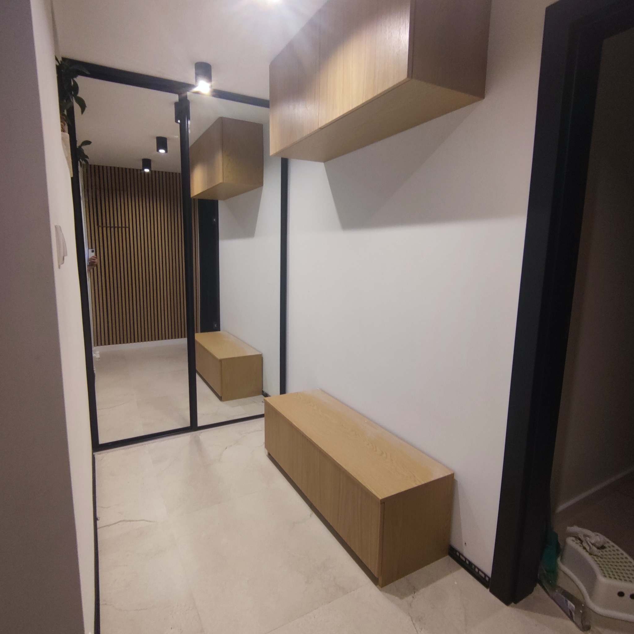 Nowoczesny przedpokój z drewnianymi szafkami wiszącymi i siedziskiem, odbicie w lustrze ukazuje ścianę z drewnianych paneli.