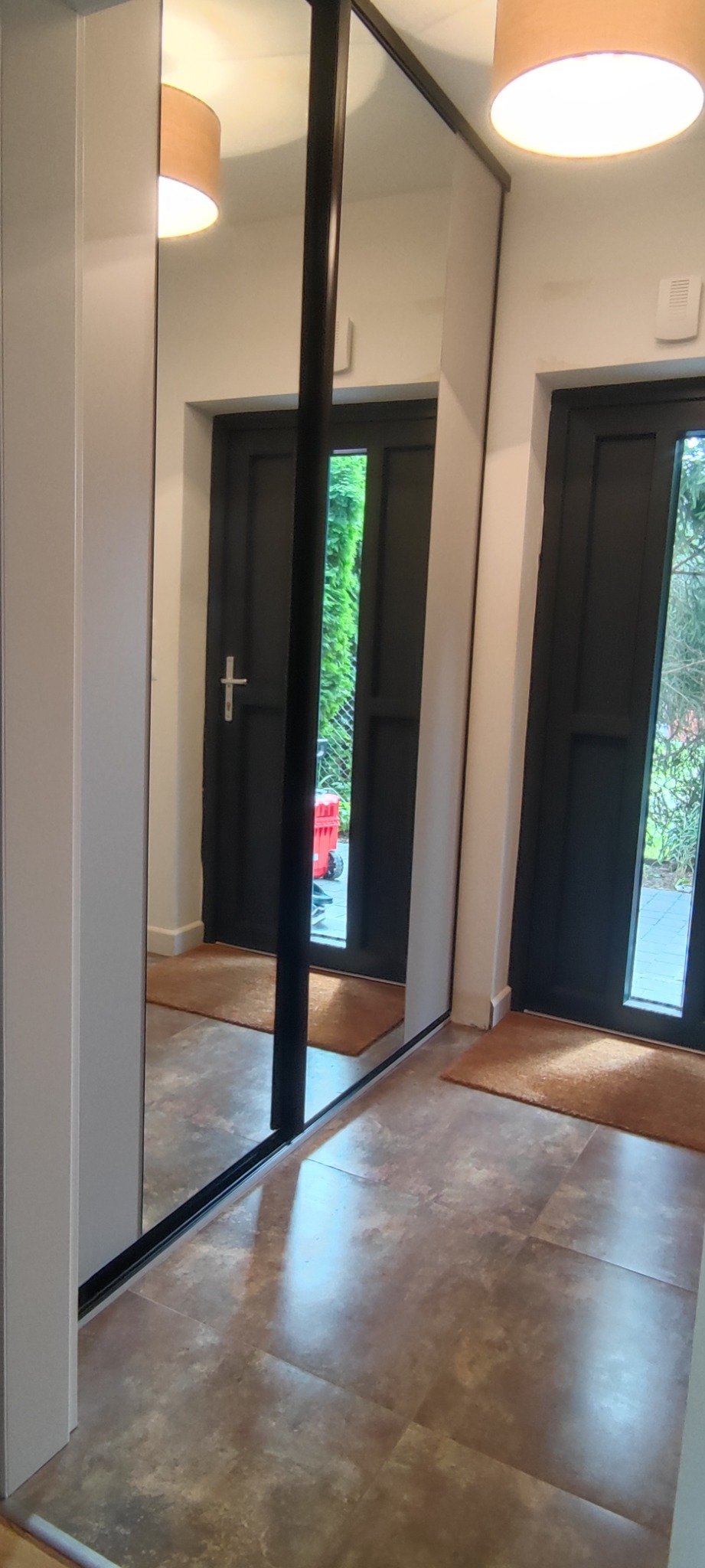 Biała szafa przesuwna z lustrzanymi drzwiami, czarne obramowanie, widok na drzwi wejściowe i otoczenie za nimi odbijające się w lustrze.