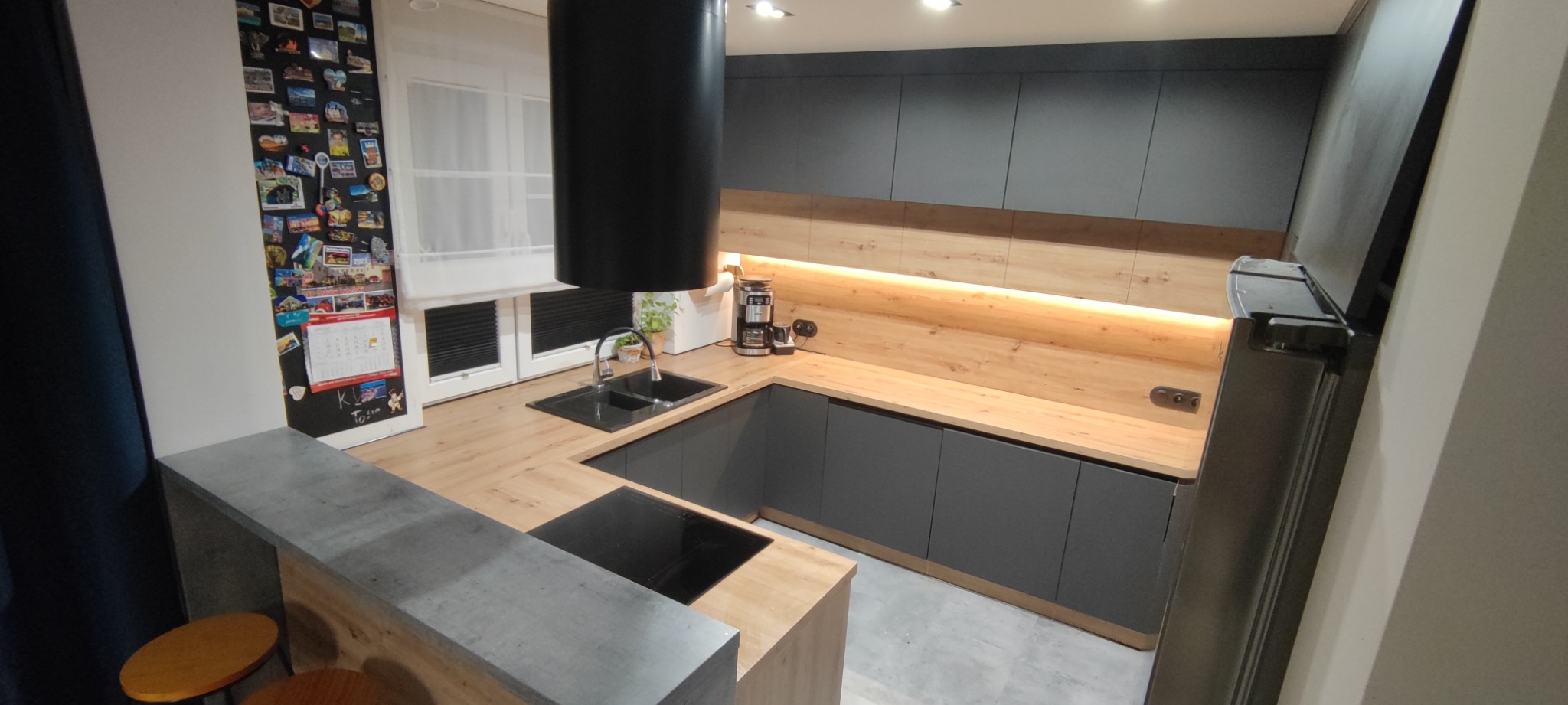 Nowoczesna kuchnia z szarymi szafkami, drewnianymi blatami i czarnym okapem cylindrycznym. Na ścianie lodówka z magnesami z podróży i kalendarzem.
