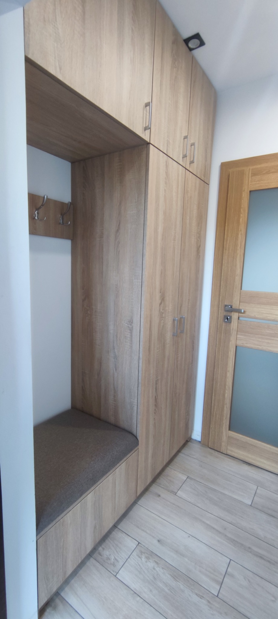 Zabudowa przedpokoju z siedziskiem i wieszakami, wykończona w okleinie drewnopodobnej, zintegrowana z szafą sięgającą sufitu.