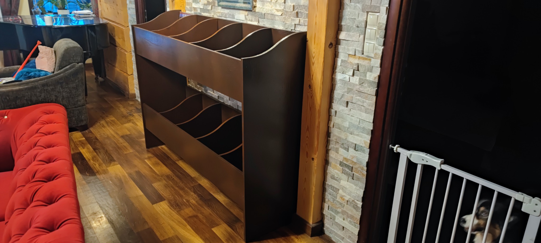 Brązowy regał na książki z półkami o falistym kształcie, ustawiony w salonie z parkietem, czerwoną sofą i elementami dekoracyjnymi z kamienia, za białą barierką widoczny pies.