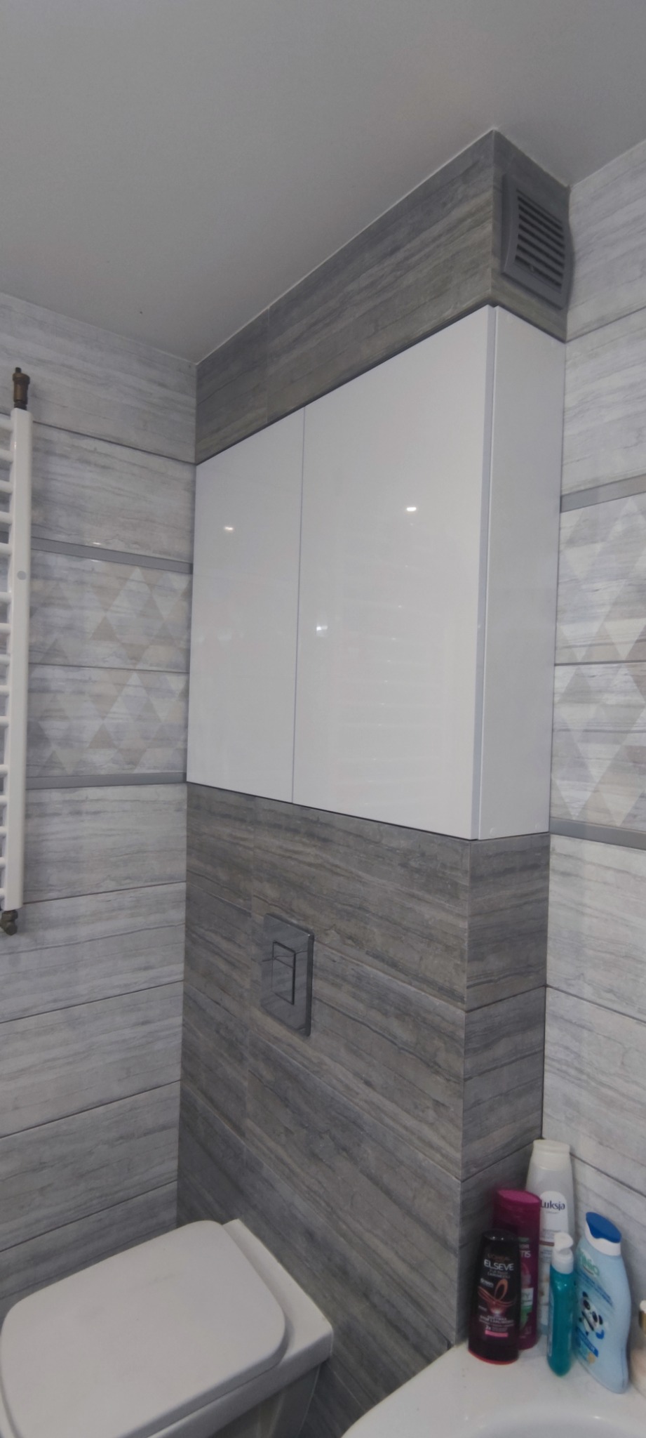 Nowoczesna łazienka z białą szafką wiszącą nad toaletą, szare płytki imitujące drewno, i widocznym wentylatorem w rogu.