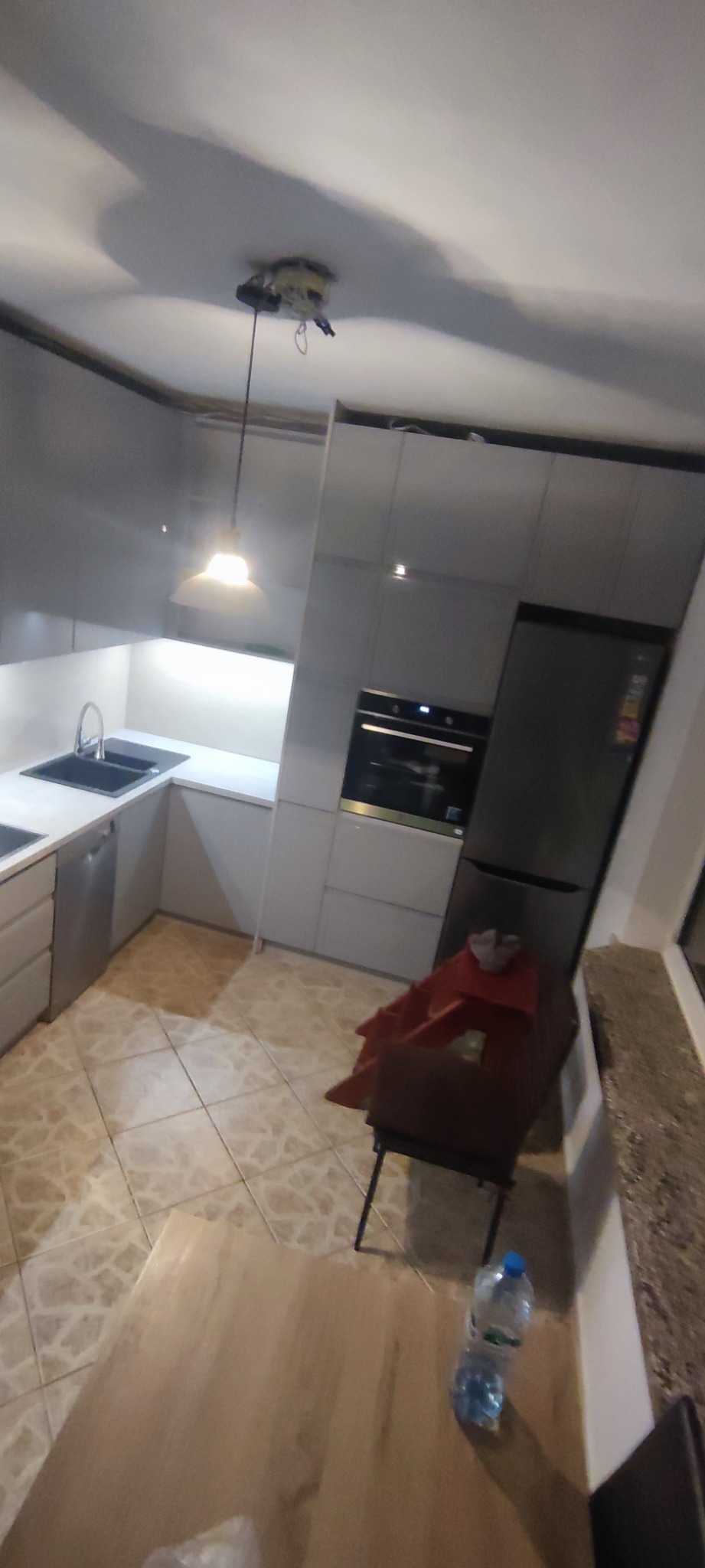 Szara, nowoczesna kuchnia na wymiar z oświetleniem podszafkowym w Lubartowie, widok na zlewozmywak, płytę grzewczą i lodówkę, z niedokończonym montażem lampy sufitowej.