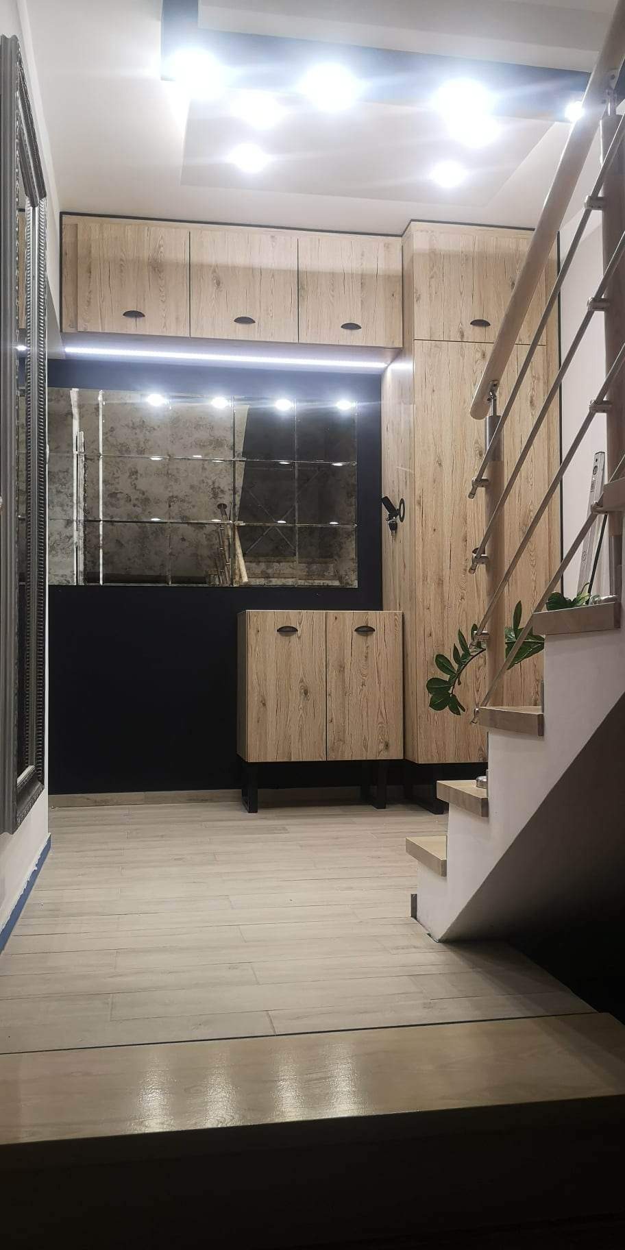 Przedpokój z zabudową z drewna w odcieniach beżu i brązu, lustro w ozdobnej ramie, schody z drewnianymi stopniami, oświetlenie LED podszafkowe.