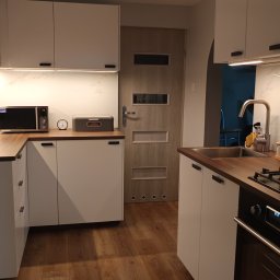 NSDESIGN.PL - Nowoczesna kuchnia z białymi szafkami, drewnianym blatem i podłogą w odcieniach brązu, oświetlona ciepłym światłem, z widocznym piekarnikiem i kuchenką gazową.