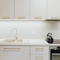 NSDESIGN.PL - Jasna kuchnia z beżowym zlewem i złotymi uchwytami, czajnik na płycie indukcyjnej, minimalistyczny design.