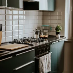 Nowoczesna kuchnia w odcieniach zieleni z białymi płytkami, czarnym blatem i stalową kawiarką na płycie gazowej.