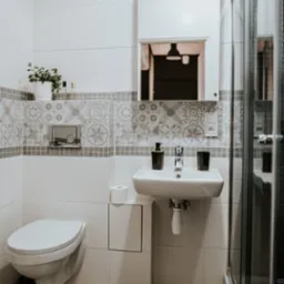 Biała łazienka z umywalką, toaletą i kabiną prysznicową, wykończona płytkami z geometrycznym wzorem, z akcentami w postaci czarnych dozowników i zielonej rośliny w doniczce.