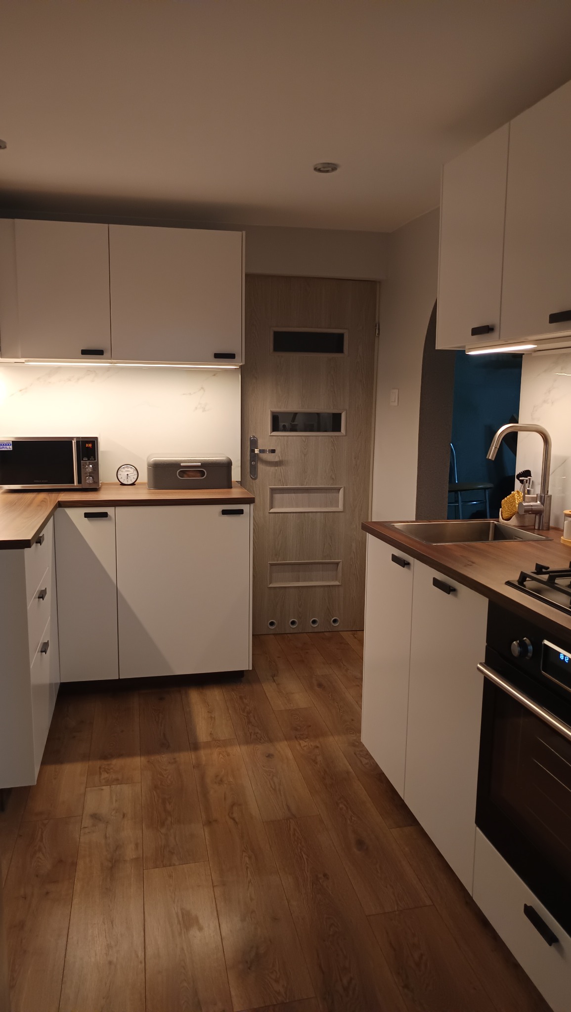 Nowoczesna kuchnia z białymi szafkami, drewnianym blatem i podłogą w odcieniach brązu, oświetlona ciepłym światłem, z widocznym piekarnikiem i kuchenką gazową.