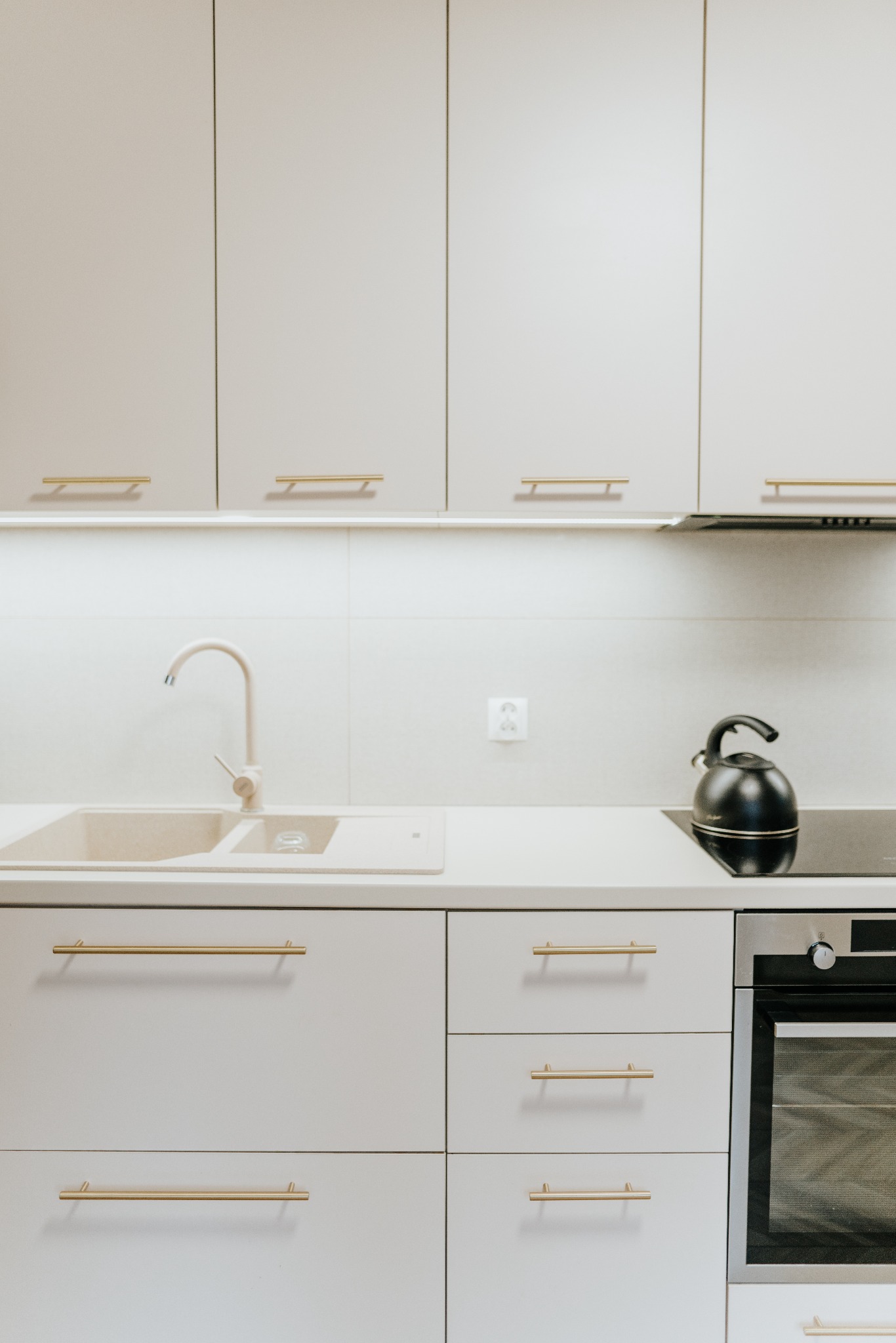 Jasna kuchnia z beżowym zlewem i złotymi uchwytami, czajnik na płycie indukcyjnej, minimalistyczny design.