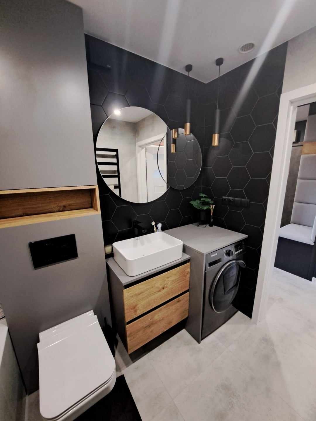 Nowoczesna łazienka z czarnymi heksagonalnymi płytkami, okrągłym lustrem, białą umywalką nablatową na drewnianej szafce, szarą zabudową WC i pralką obok umywalki.