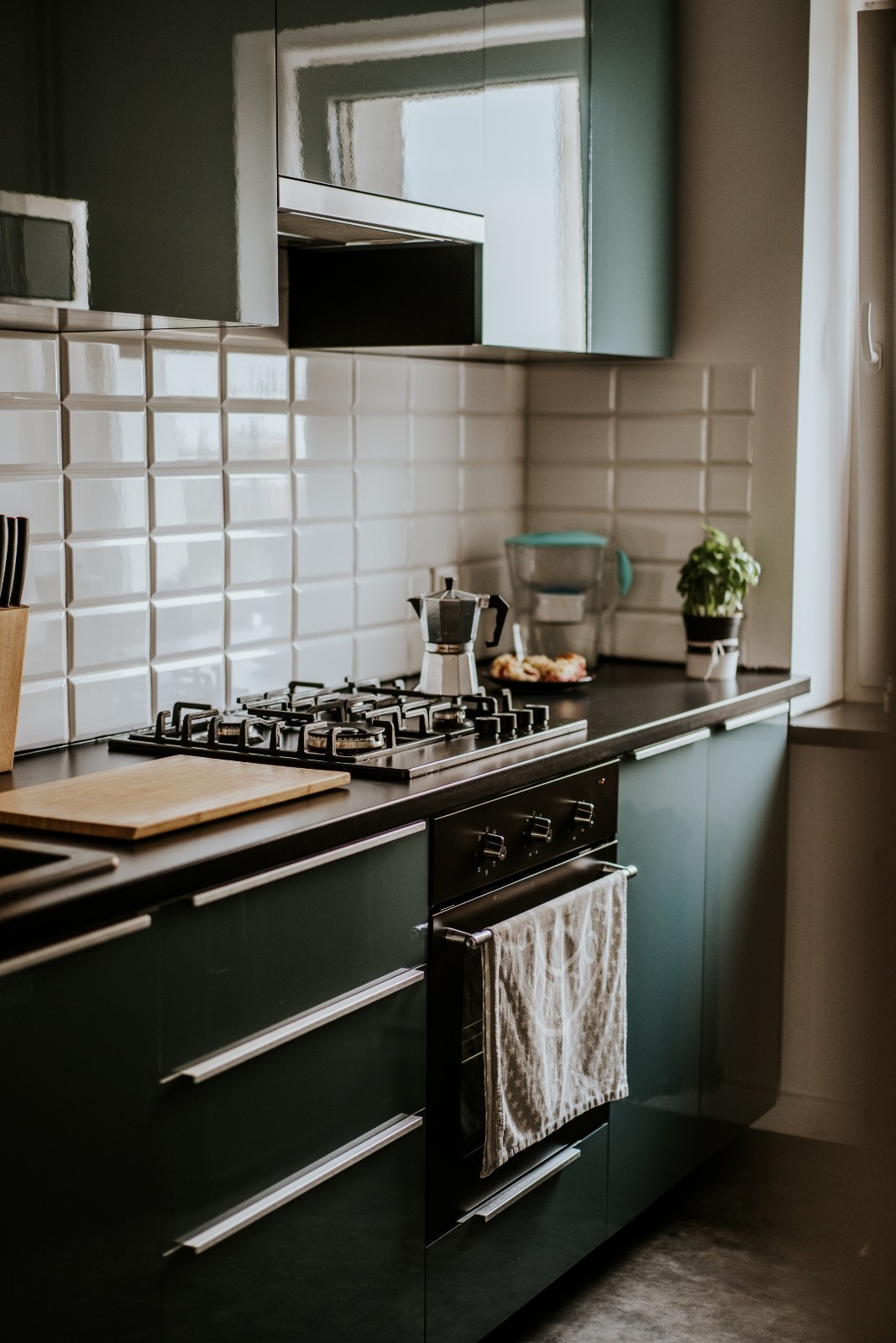 Nowoczesna kuchnia w odcieniach zieleni z białymi płytkami, czarnym blatem i stalową kawiarką na płycie gazowej.