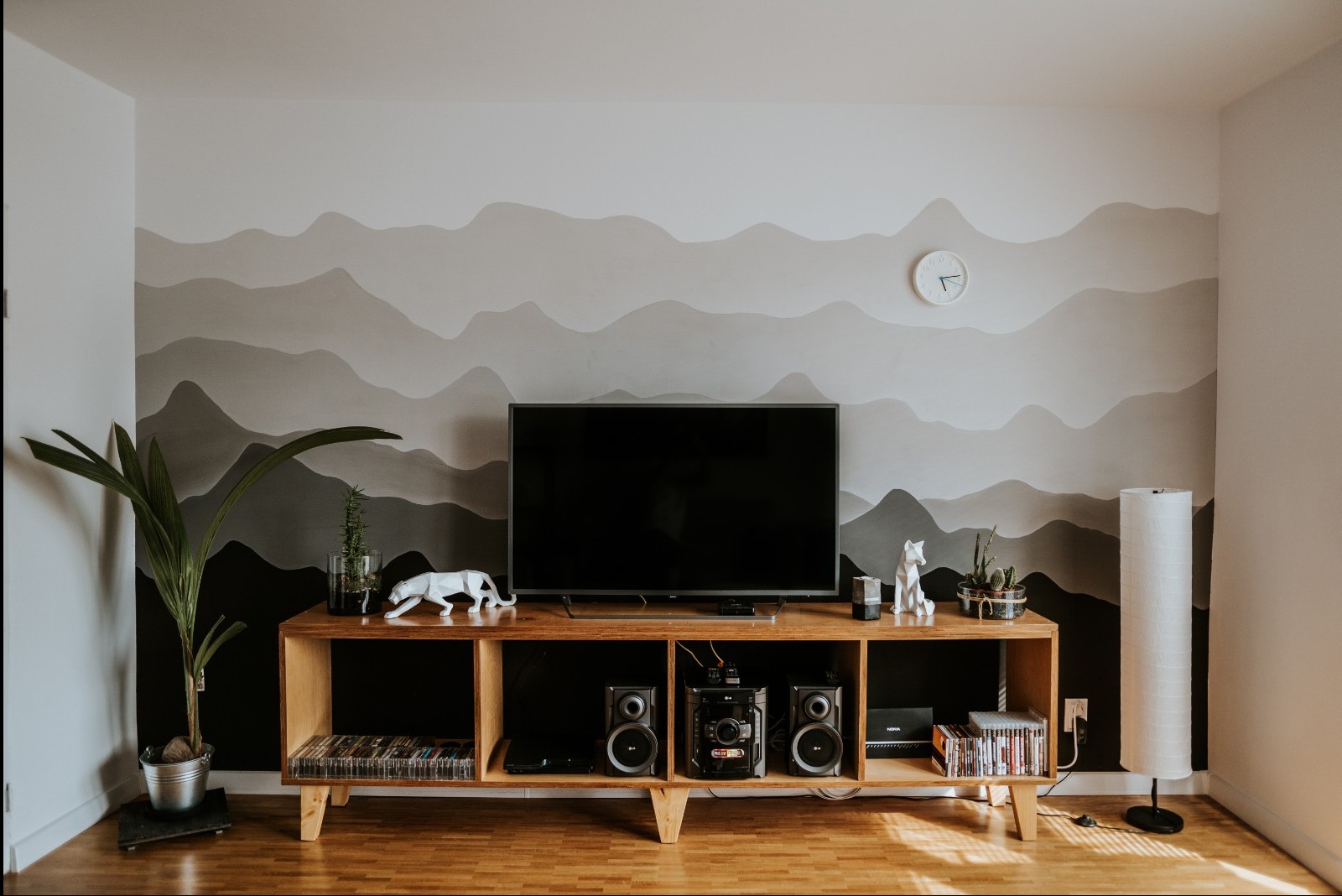 Salon z drewnianą szafką RTV, telewizorem, dekoracyjną ścianą w odcieniach szarości imitującą góry, roślinami doniczkowymi i minimalistyczną lampą podłogową.