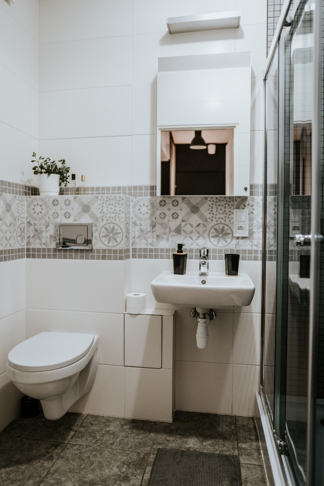 Biała łazienka z umywalką, toaletą i kabiną prysznicową, wykończona płytkami z geometrycznym wzorem, z akcentami w postaci czarnych dozowników i zielonej rośliny w doniczce.
