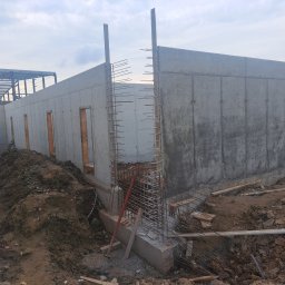 WBWP SPÓŁKA Z OGRANICZONĄ ODPOWIEDZIALNOŚCIĄ - Surowe ściany z betonu architektonicznego z otworami okiennymi i zbrojeniami, widoczne na placu budowy z hałdą ziemi.