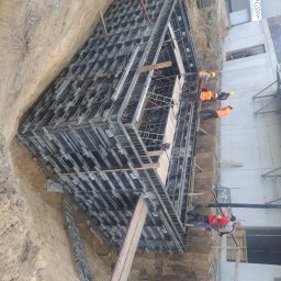 WBWP SPÓŁKA Z OGRANICZONĄ ODPOWIEDZIALNOŚCIĄ - Budowa fundamentów: ekipa budowlana w żółtych kaskach i pomarańczowych kamizelkach pracuje przy szalunku fundamentu w wykopie, widoczny betonowy mur w tle.