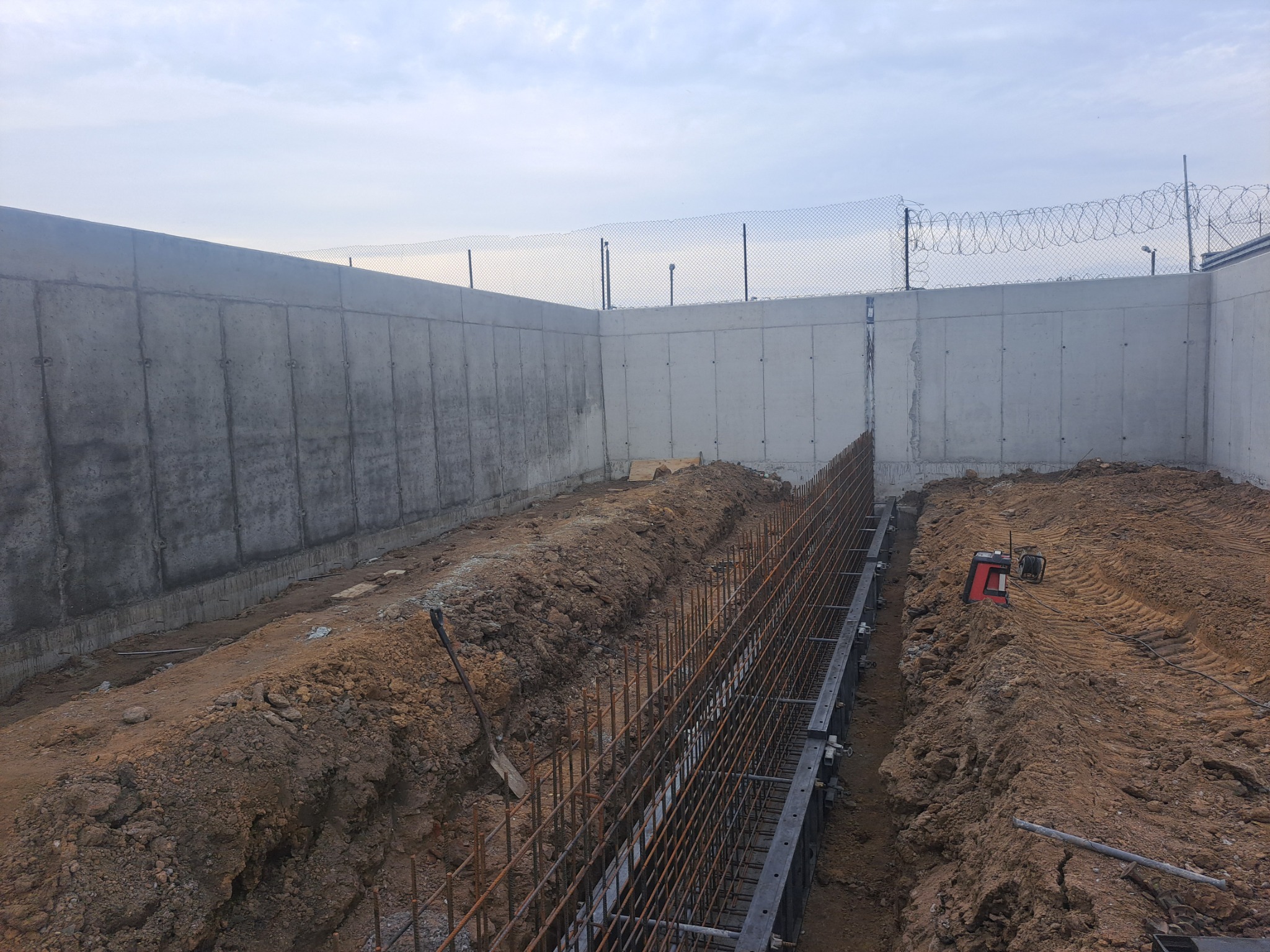 Przygotowanie fundamentów: wykop z szalunkiem i zbrojeniem stalowym, otoczony ziemią, z widocznym betonowym murem oporowym w tle pod błękitnym niebem.
