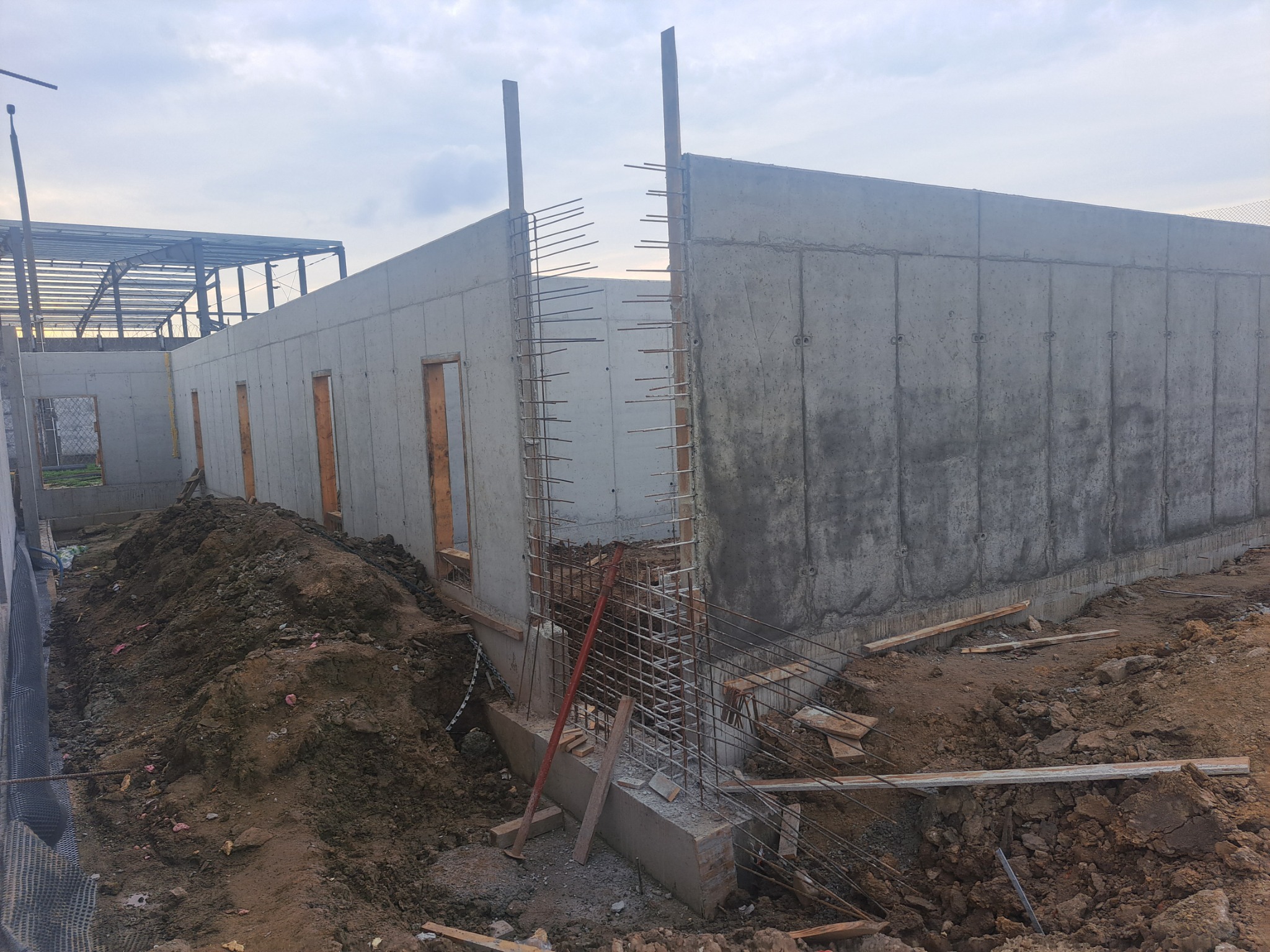 Surowe ściany z betonu architektonicznego z otworami okiennymi i zbrojeniami, widoczne na placu budowy z hałdą ziemi.