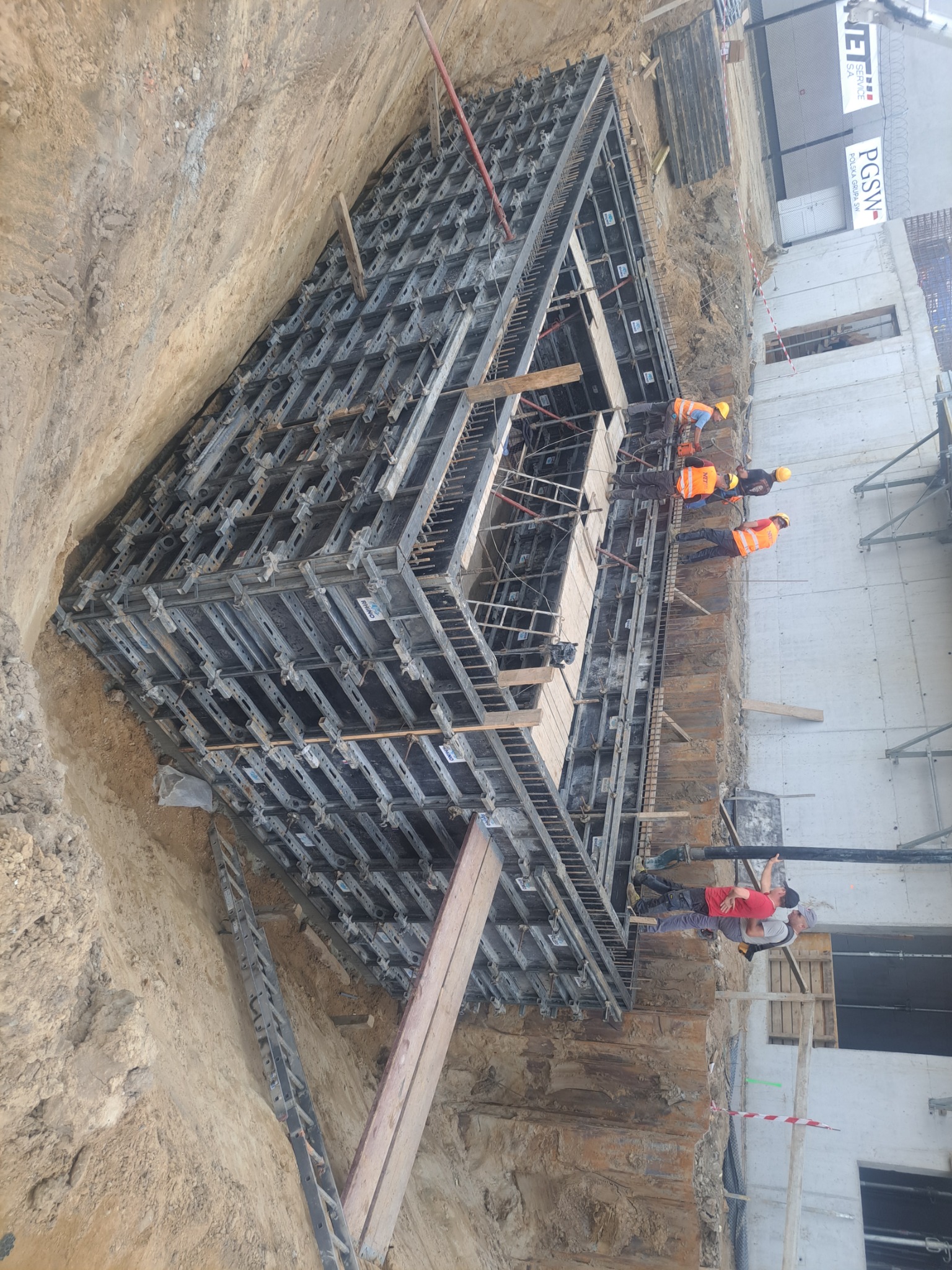 Budowa fundamentów: ekipa budowlana w żółtych kaskach i pomarańczowych kamizelkach pracuje przy szalunku fundamentu w wykopie, widoczny betonowy mur w tle.