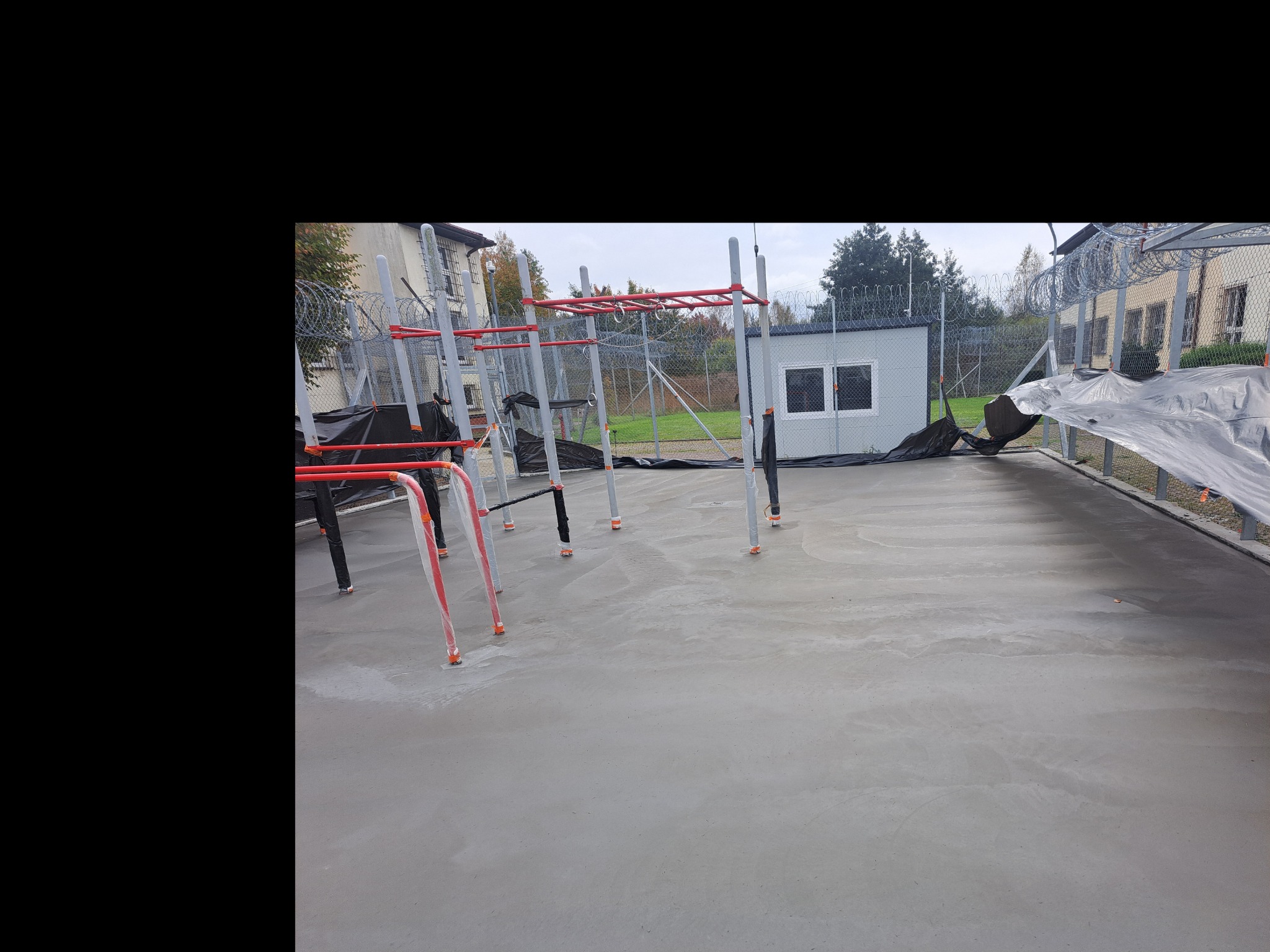 Świeżo wylany betonowy plac zabaw z zamontowanymi elementami konstrukcji do ćwiczeń, zabezpieczonymi folią, otoczony ogrodzeniem z drutem kolczastym.