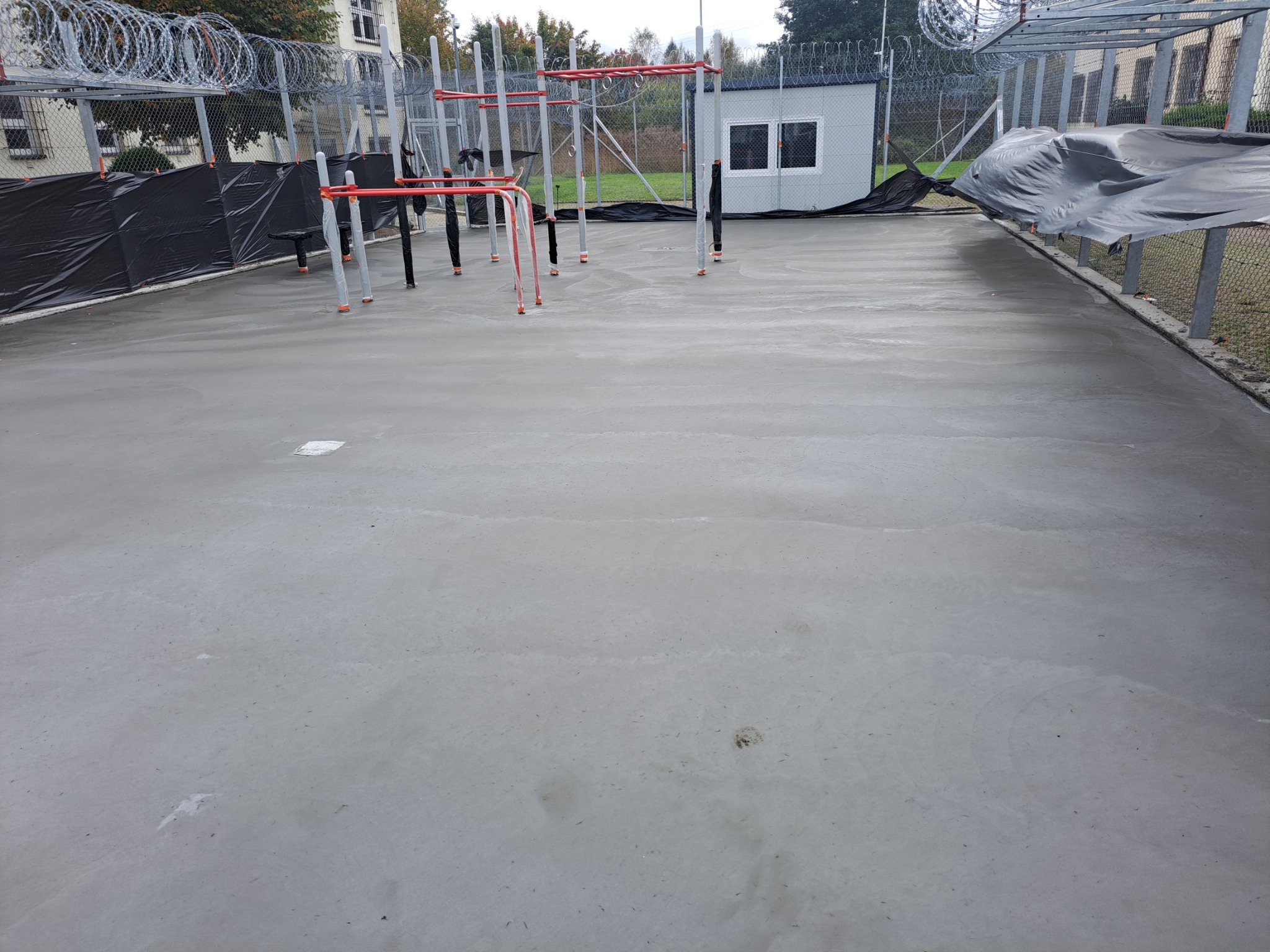 Świeżo wylana, gładka betonowa posadzka na zewnątrz z zamontowanymi elementami metalowej konstrukcji placu zabaw lub siłowni zewnętrznej, otoczona ogrodzeniem z drutem kolczastym.