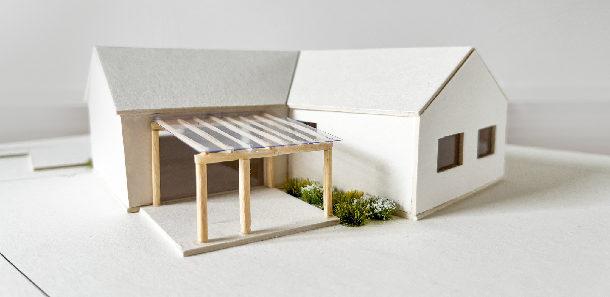Biały, minimalistyczny model domu z przeszklonym zadaszeniem nad wejściem, otoczony drobną zielenią. Precyzyjne wykonanie, neutralne tło.