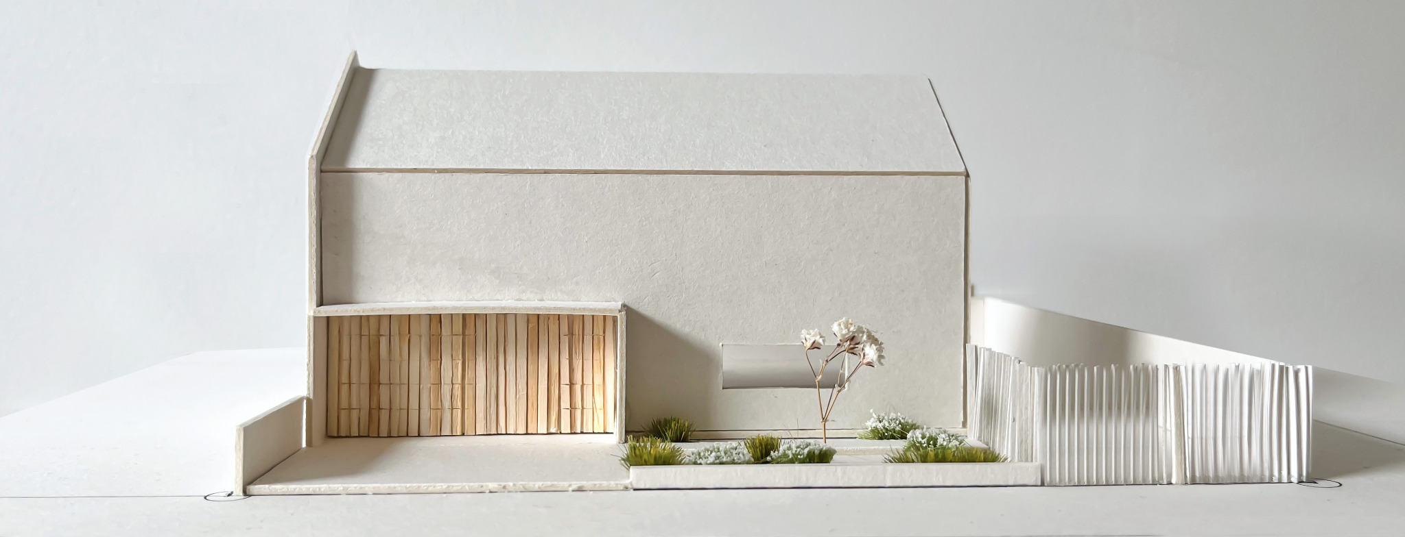 Architektoniczna makieta domu z jasnego kartonu, ukazująca minimalistyczną bryłę z drewnianymi akcentami i elementami zieleni w otoczeniu.