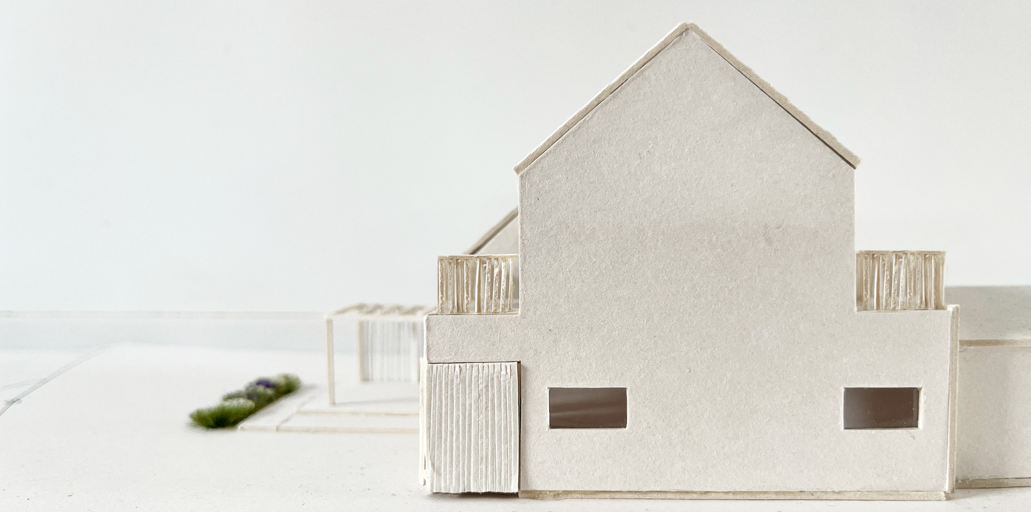 Architektoniczna makieta domu z tektury, ukazująca detale elewacji, balkonu i otoczenia w minimalistycznym stylu na jasnym tle.