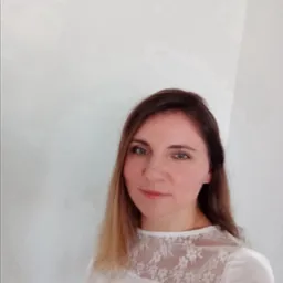 Portret kobiety z długimi brązowymi włosami i białej bluzce z koronkowym wykończeniem pod szyją, na tle jasnej ściany.