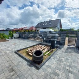 DaMaR Usługi koparką - Ułożona szara kostka brukowa na posesji z elementami dekoracyjnymi: opona i drewno, w tle brama wjazdowa i zaparkowany bus firmy.