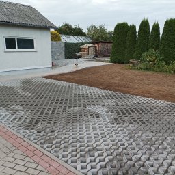 DaMaR Usługi koparką - Ułożona szara ażurowa nawierzchnia z kostki brukowej przechodzi w przygotowany teren pod trawnik, w tle budynek gospodarczy, wysokie tuje i mały pies.