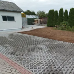 Ułożona szara ażurowa nawierzchnia z kostki brukowej przechodzi w przygotowany teren pod trawnik, w tle budynek gospodarczy, wysokie tuje i mały pies.