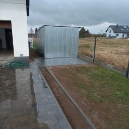 DaMaR Usługi koparką - Ułożona kostka brukowa wzdłuż domu, prowadząca do metalowego garażu. Widoczny trawnik z obrzeżami i nowa kostka pod garażem. Całość w pochmurny dzień.