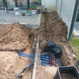 DaMaR Usługi koparką - Wykop z niebieskimi rurami instalacji nawadniającej w ziemi, obok budynku i ogrodzenia panelowego. Widoczny sterownik nawadniania na ścianie budynku.