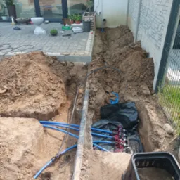 Wykop z niebieskimi rurami instalacji nawadniającej w ziemi, obok budynku i ogrodzenia panelowego. Widoczny sterownik nawadniania na ścianie budynku.