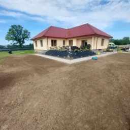 Przygotowany teren pod trawnik z rolki przed domem jednorodzinnym. Widoczna świeżo nawieziona ziemia i obrzeża. W tle dom z czerwonym dachem i fragment ogrodzenia.
