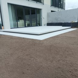 DaMaR Usługi koparką - Nowoczesny taras z jasnych płyt na przygotowanym podłożu z ziemi ogrodowej, przylegający do budynku z dużymi oknami przesuwnymi i minimalistycznym ogrodzeniem.