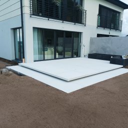 DaMaR Usługi koparką - Nowoczesny taras z jasnych płyt na przygotowanym podłożu, widok na budynek z dużymi oknami i balkonem z czarną balustradą, ziemia ogrodowa w tle.