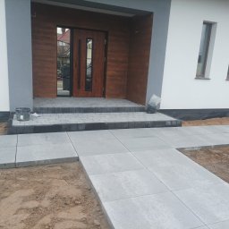 DaMaR Usługi koparką - Nowoczesne wejście do domu z szarej kostki brukowej. Ułożony chodnik prowadzi do drewnianych drzwi wejściowych. Widoczne elementy elewacji i przygotowany teren.