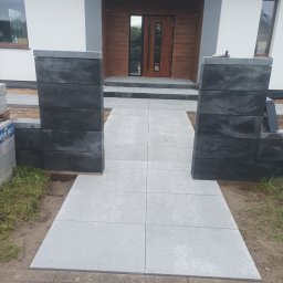 DaMaR Usługi koparką - Nowoczesna ścieżka z szarych płyt prowadząca do domu, flankowana przez ciemne, modułowe słupki. Widoczne detale wykończenia i paleta z kostką brukową.