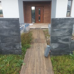 DaMaR Usługi koparką - Wejście do domu z drewnianą ścieżką i elementami ogrodzenia z szarej kostki. Widoczne materiały budowlane i skrzynka na listy. Ujęcie z perspektywy.