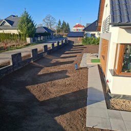 DaMaR Usługi koparką - Ułożona kostka chodnikowa przy domu, przygotowany teren pod trawnik, w tle widoczny fragment ogrodzenia z betonowych bloczków. Słoneczny dzień.